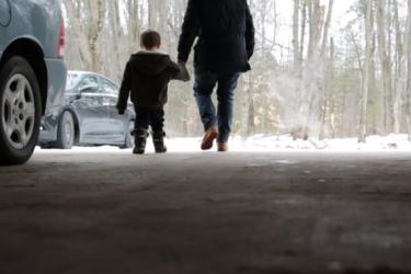 Adult holding the hand of a child seen from the back exiting a garage walking towards a snow covered forest