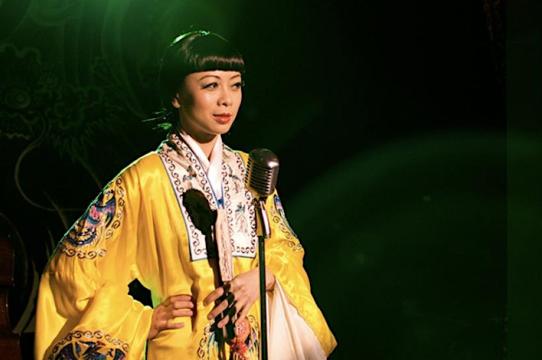 Asian woman with short bangs wearing a yellow robe standing in front of a microphone