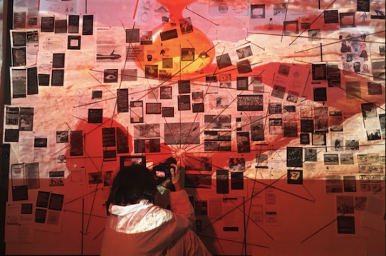 A woman takes pictures in front of artwork map with red hue.