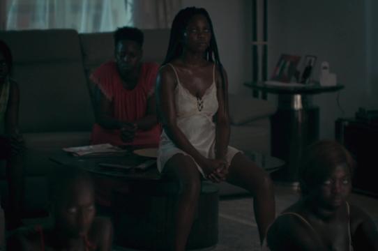 dark image of a black woman wearing a white slip sitting on a coffee table with a black man in a red shirt behind her