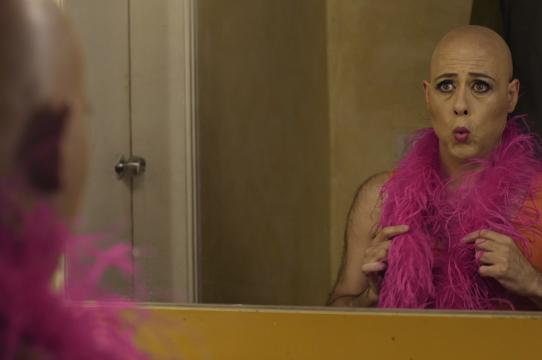 Bald man wearing a pink boa looking at himself in the mirror