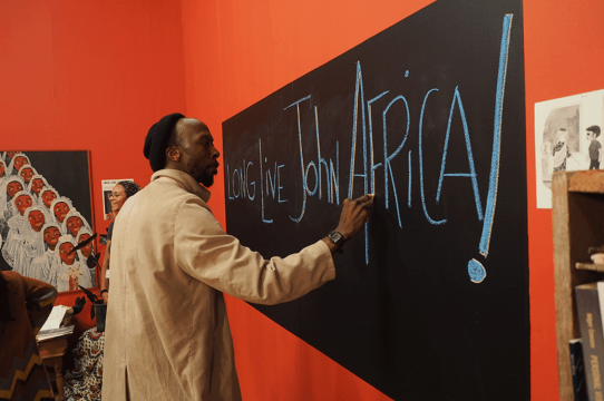Black Man in a trenchcoat writing on a chalkboard "Long Live John Africa!"